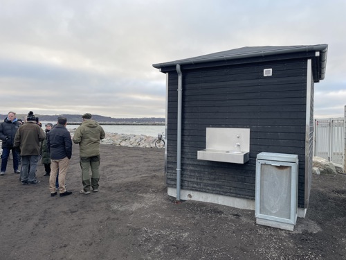 Toiletbygningen set fra siden, hvor fremmødte kan ses i baggrunden